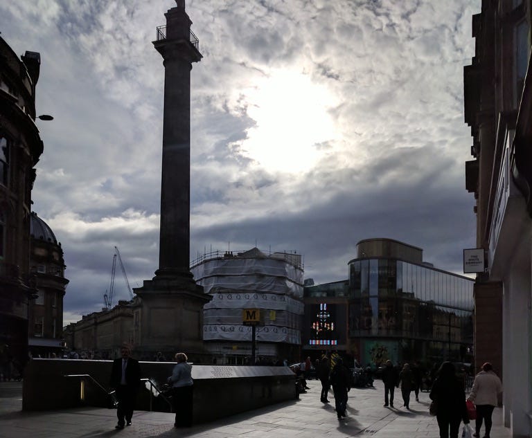 Newcastle City Centre near Monument. It is a bright and sunny day. It is not very busy, surrounded by buildings both old and new.