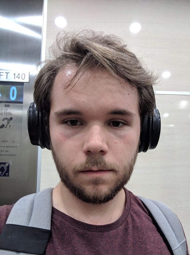 A masculine non-binary person standing in an elevator. They do not look well. There is a distant look in their eyes.