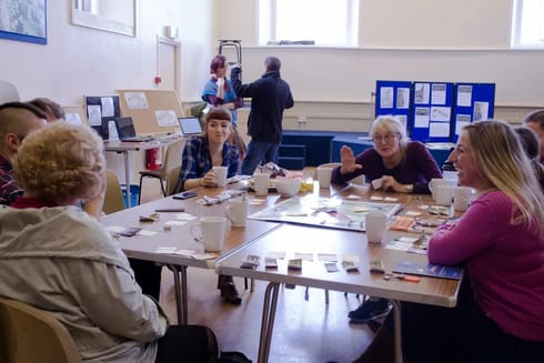 A group of white people around a table in discussion about a board game. 