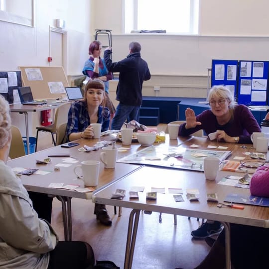 A group of white people around a table in discussion about a board game. 